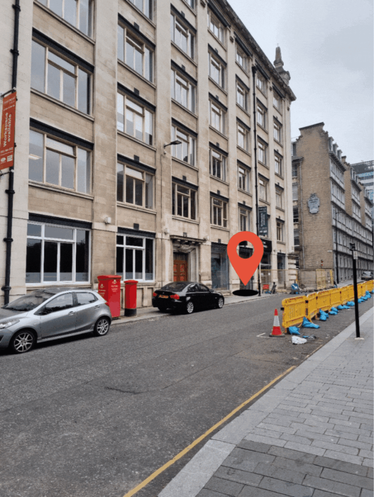 A multi-story building with parked cars along the road. A red location pin marks a specific entry door among several, often used for weekly meetings. Orange barricades line the right side of the street.