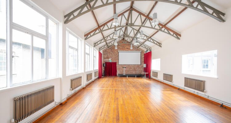 A spacious, well-lit room with a vaulted ceiling, wooden floor, large windows, radiators, and a brick wall featuring a screen and projector stand—perfect for hosting your weekly meetings in style.