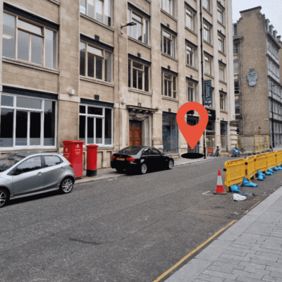 A multi-story building with parked cars along the road. A red location pin marks a specific entry door among several, often used for weekly meetings. Orange barricades line the right side of the street. A multi-story building with parked cars along the road. A red location pin marks a specific entry door among several, often used for weekly meetings. Orange barricades line the right side of the street.
